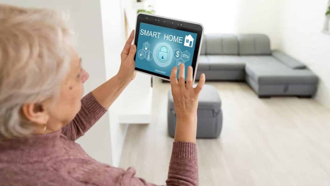An older woman holds a tablet device that says "Smart Home" on the screen. The woman is standing in her living room, pushing buttons on the tablet.