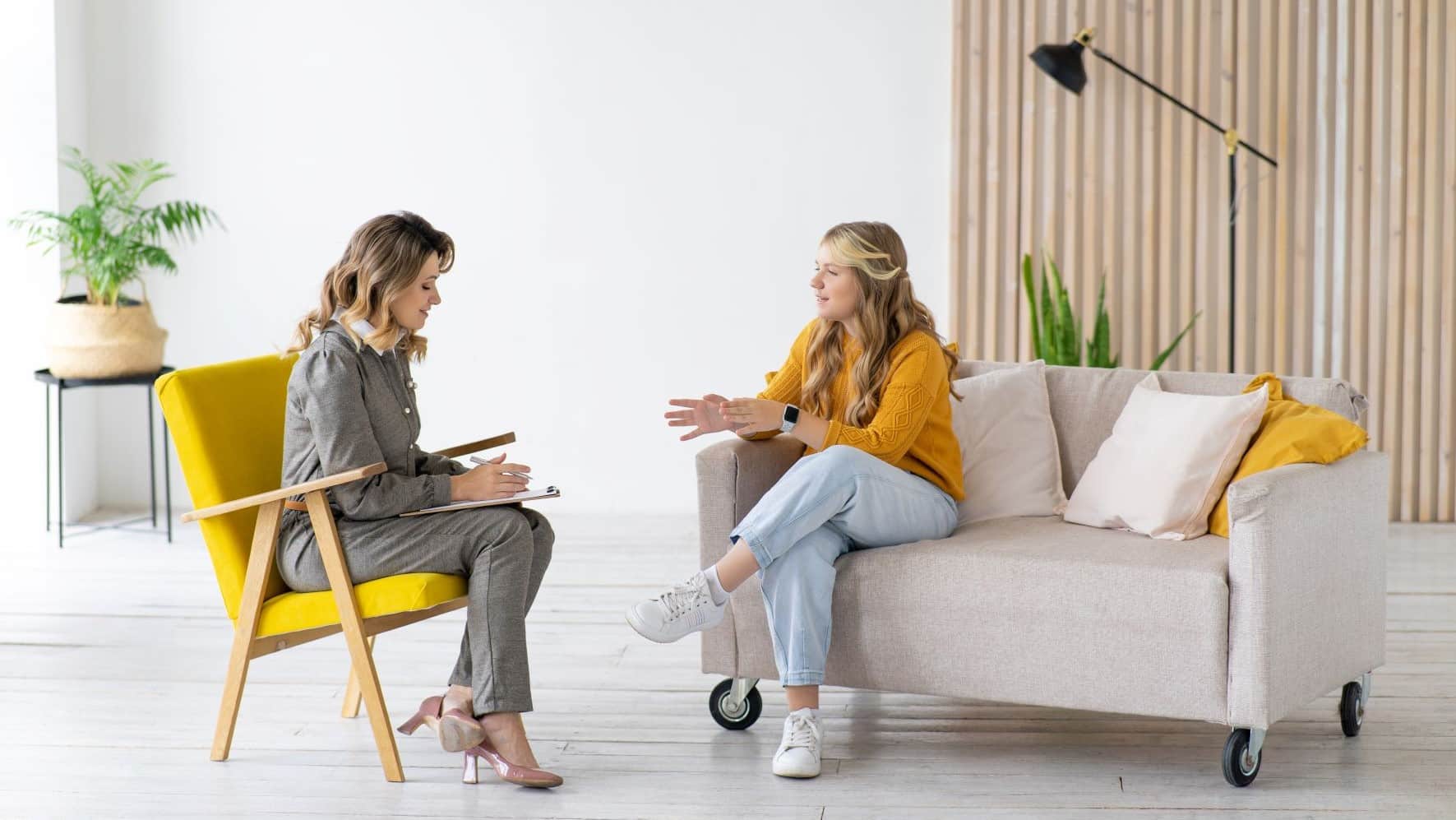 Young adult woman in an orange sweater sitting on a white sofa talking to a woman therapist with a clipboard. The therapist is wearing a gray suit.