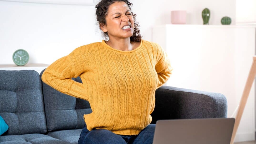 Woman sitting on a couch, holding her back and grimacing in pain.