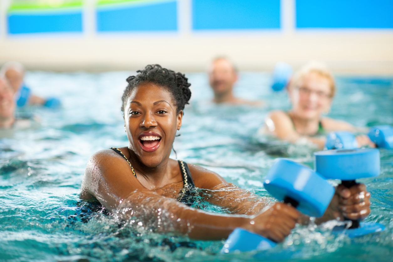 woman in pool takes exercise class, holding foam hand weights and smiling