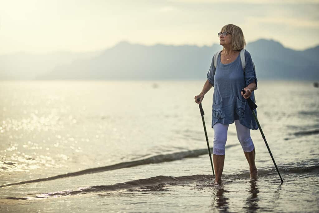 Woman walking through shallow water on the beach with walking sticks