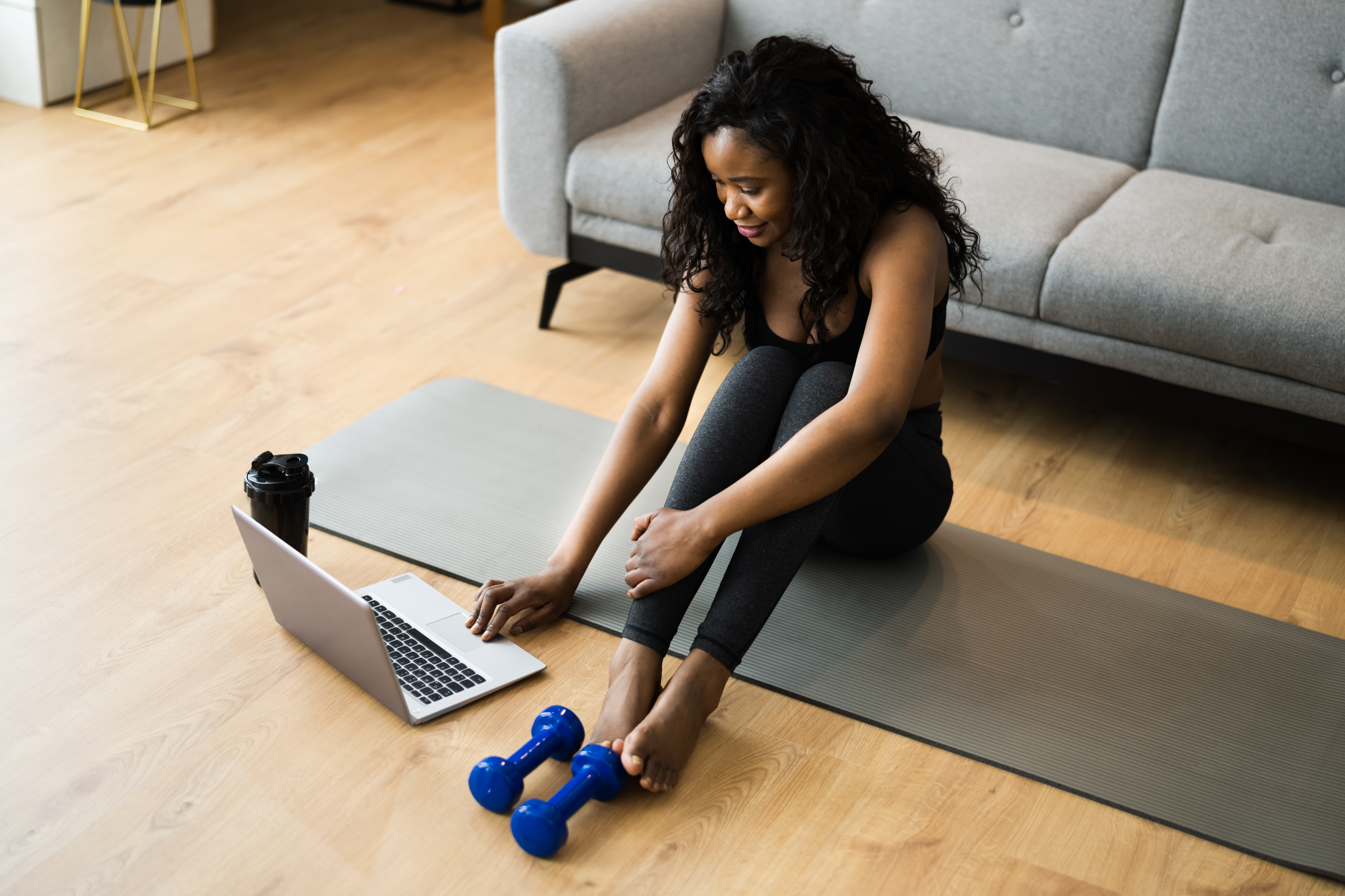 woman is seated on an exercise mat with laptop and hand weights