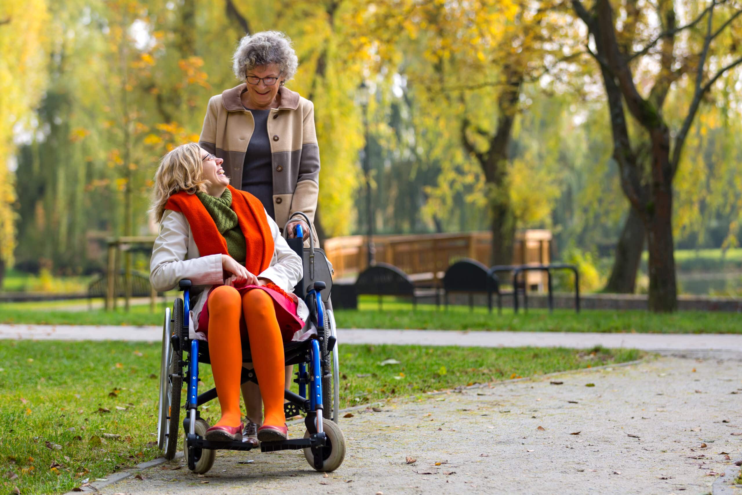 Mother pushing daughter in wheelchair in the park, smiling at one another