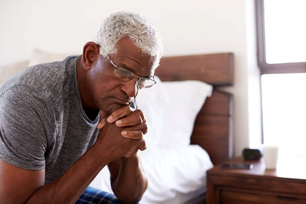 Older man sitting on edge of bed looking unhappy