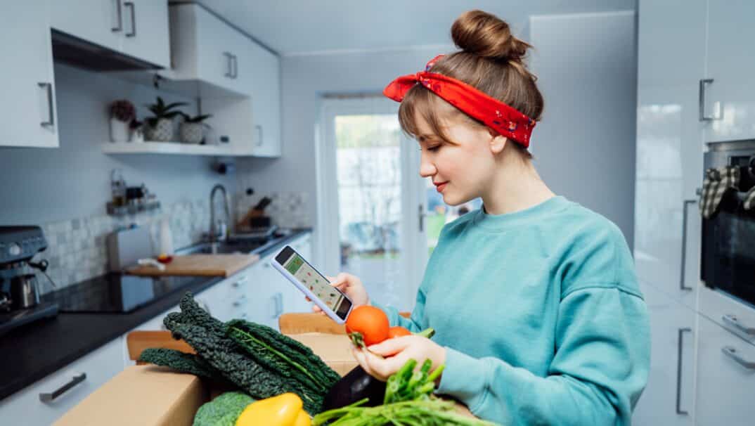 Woman in a blue sweatshirt and red headband is standing in her kitchen with a box of groceries, looking at her phone.