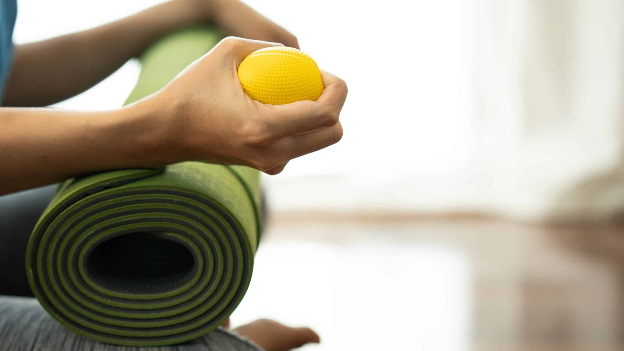 Person sitting down with a yoga mat rolled up in their arms. In their hand, they are holding a yellow yoga ball. No faces are shown in this image.