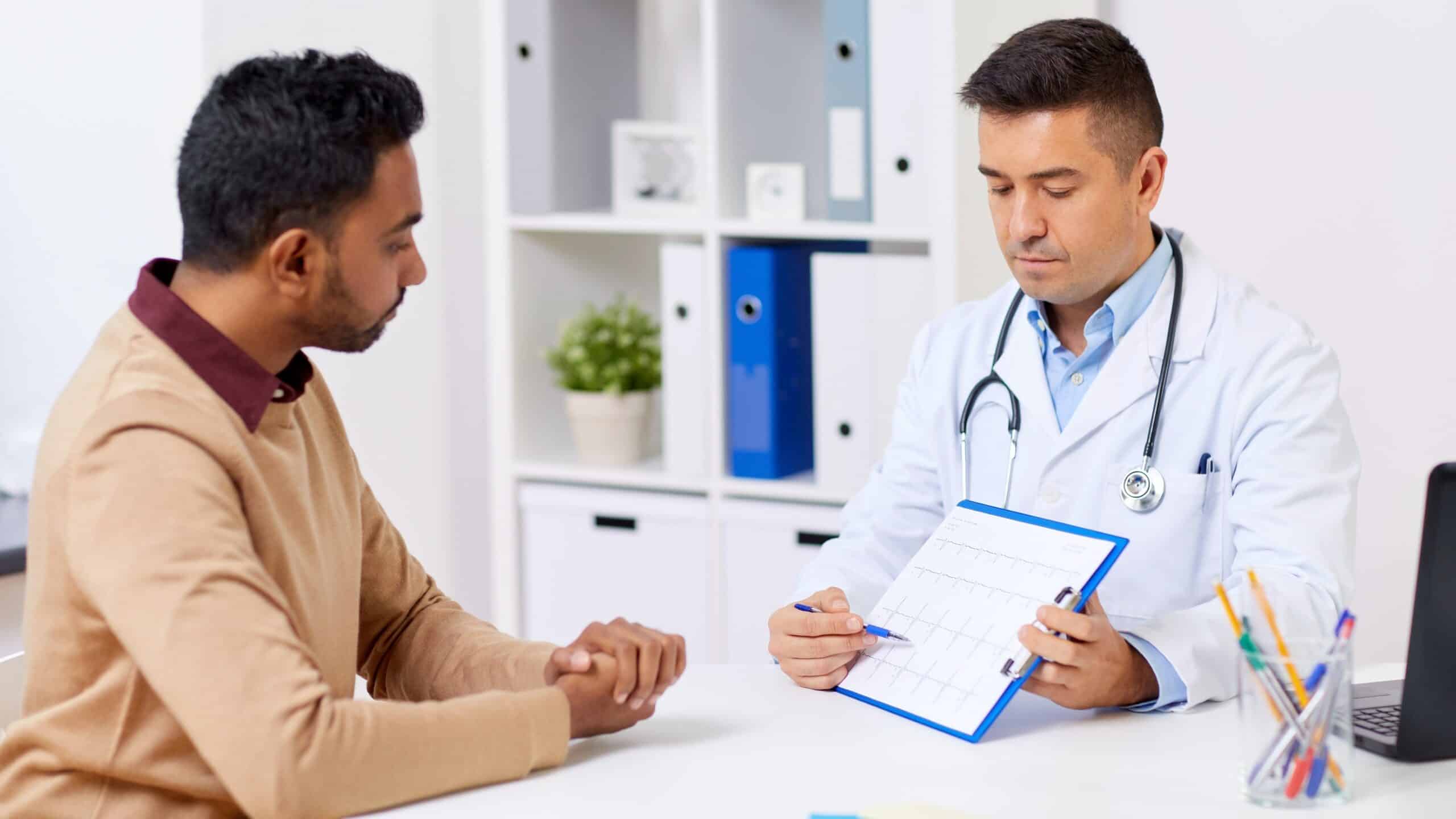Doctor showing Hispanic man something on a clipboard as they sit across a desk from one another.