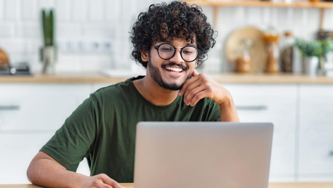 Young adult Latino man smiling while looking at his laptop.