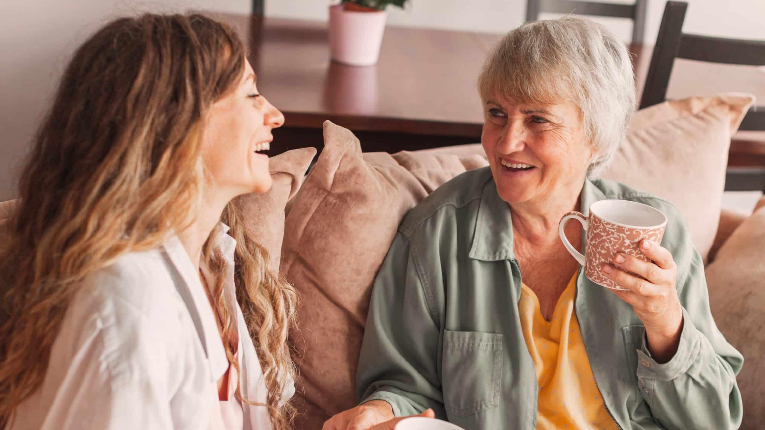 A woman with dark, long hair in conversation with her friend, a woman with short white hair. They are sitting on a couch drinking coffee together.