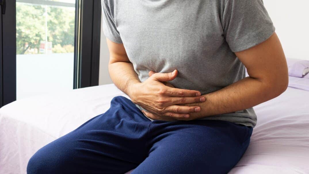 A man holding his mid section while sitting down on his bed.