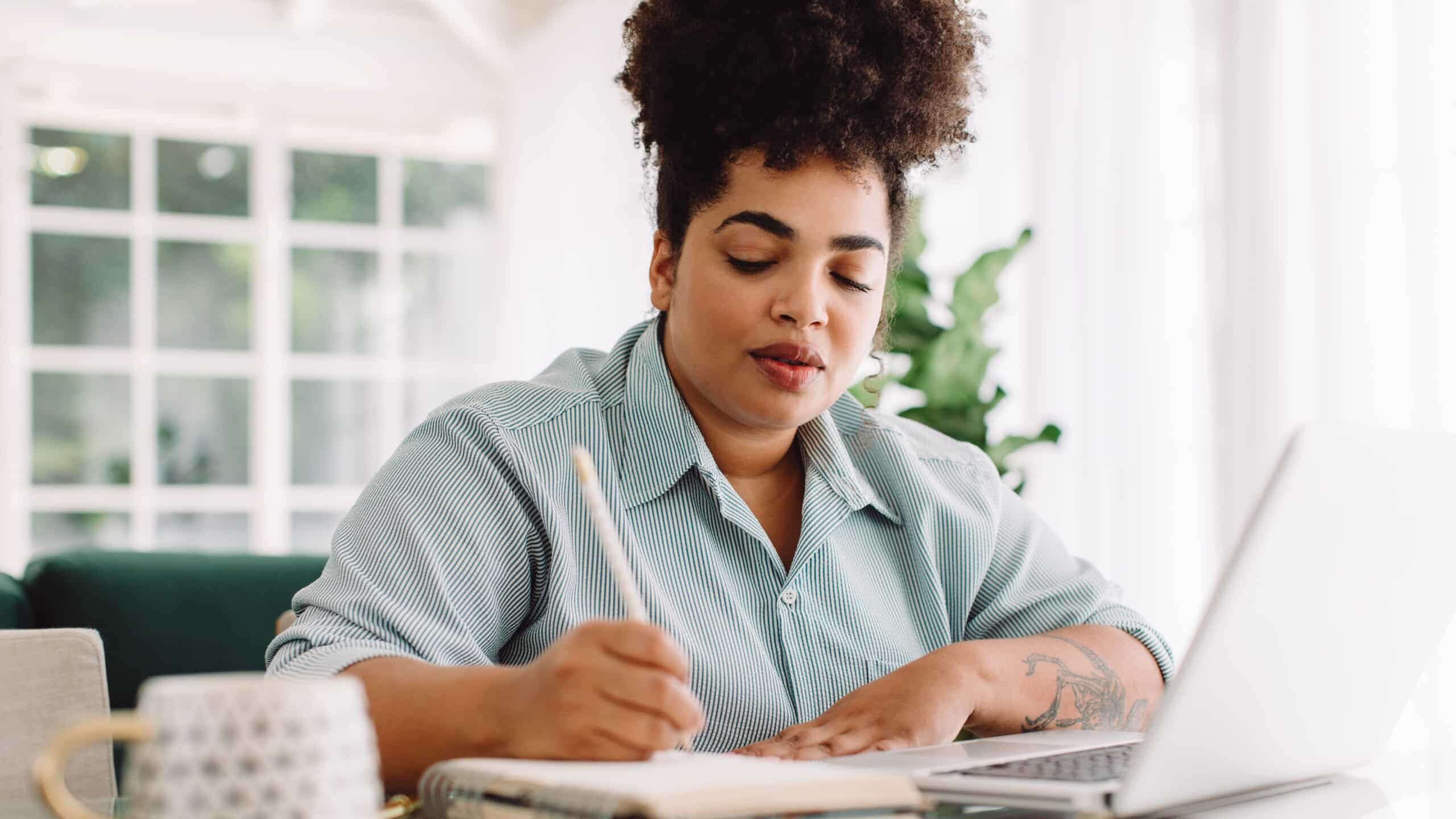 A young adult Black woman taking written notes while watching something on her laptop.