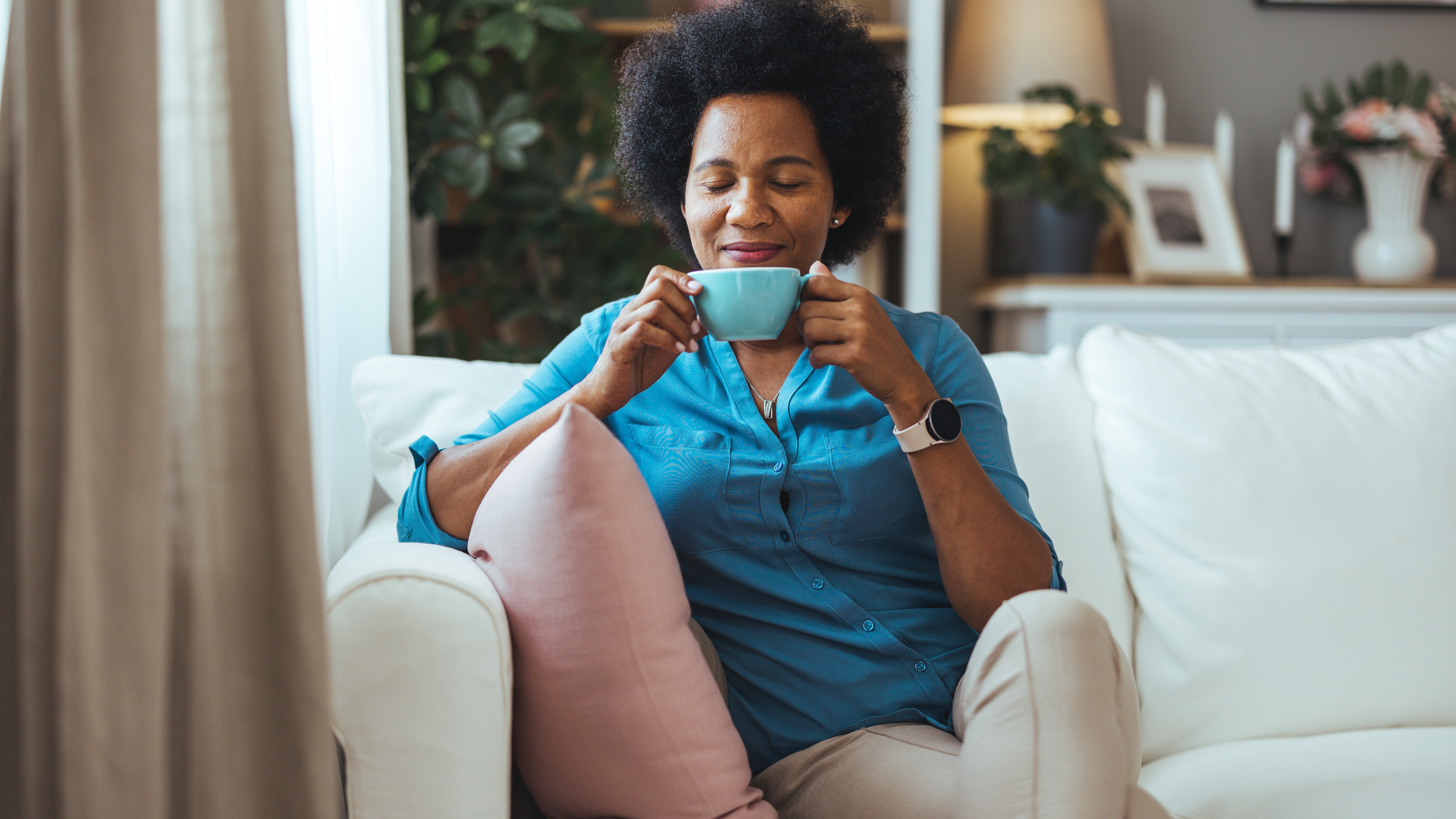 A Black woman sitting on her couch with her eyes closed. She is holding a mug of coffee.