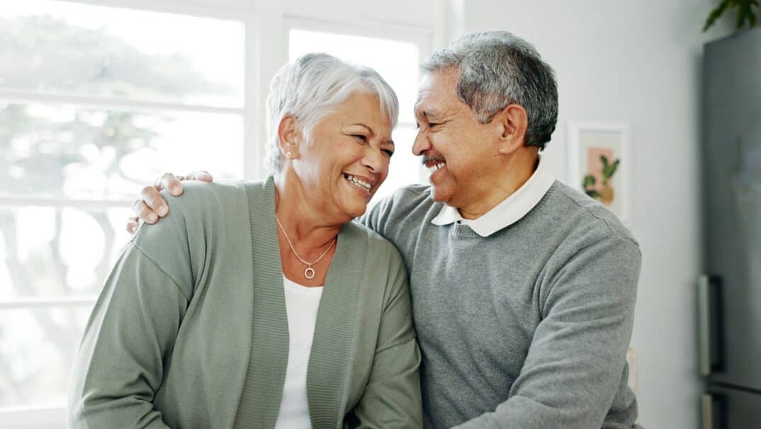 Middle aged Hispanic couple smiling at one another. The man has his arm around his wife and they are embracing side by side.