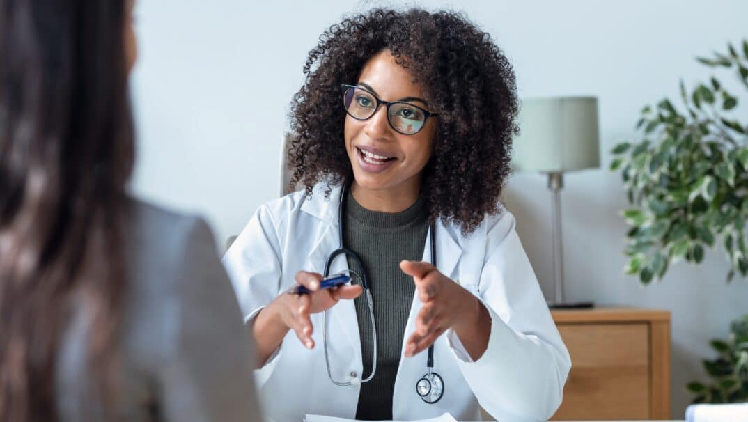 Young, Black female doctor speaking across her desk to a patient.