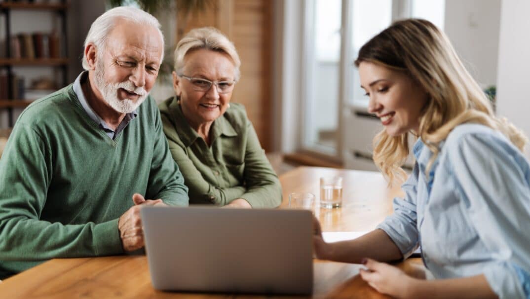 An older couple is sitting across the table from a young, female financial advisor. The financial advisor is showing the couple something on her computer.