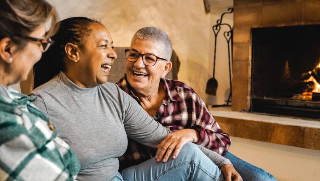 Group of older women laughing together while sitting by a lit fireplace.