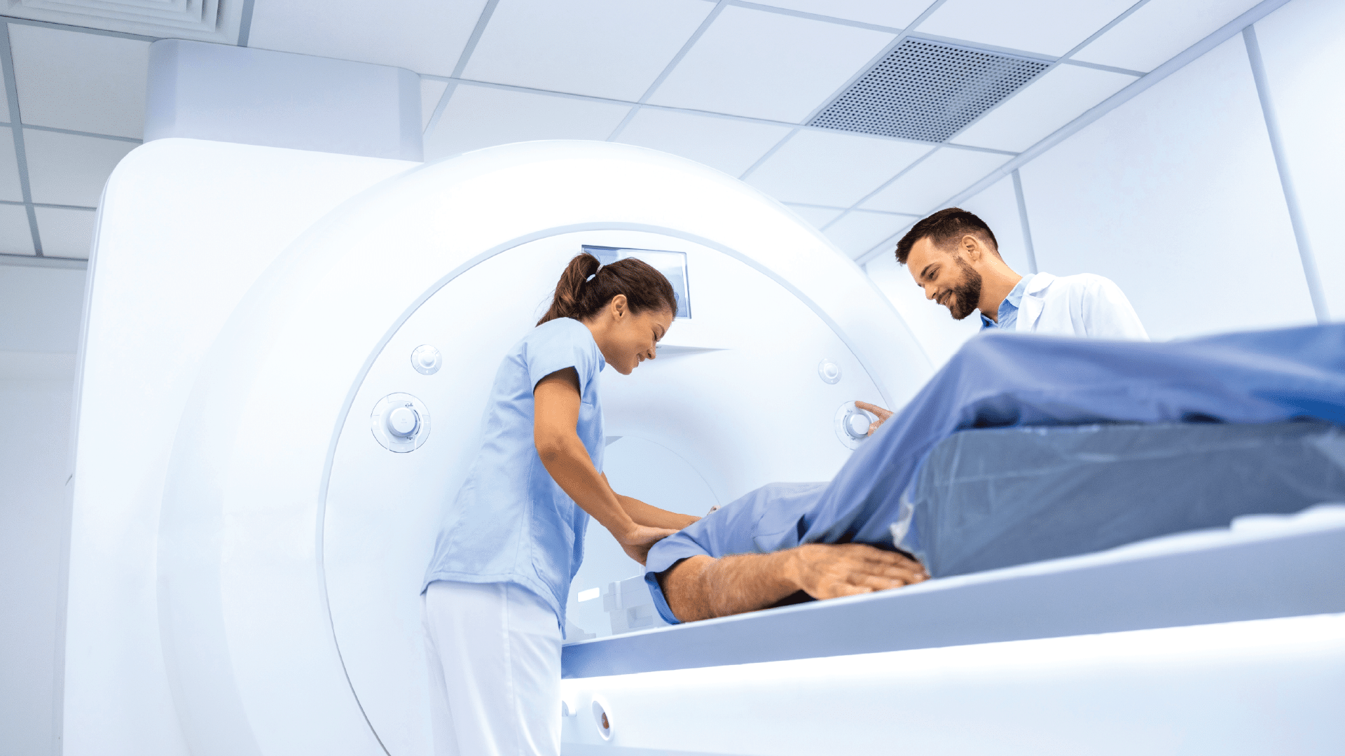 Two healthcare works support a patient entering an MRI machine.