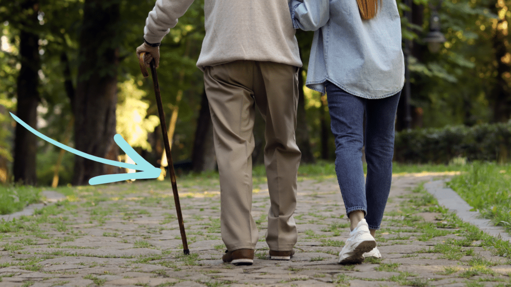 A woman and man walking on a path outside together with their backs to the camera. The man is using a cane.