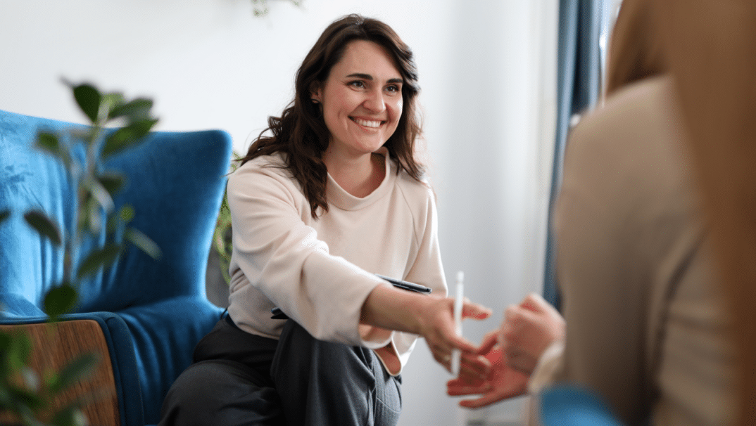 A female, brown-haired therapist shakes a clients hand. She is smiling and on a blue couch.