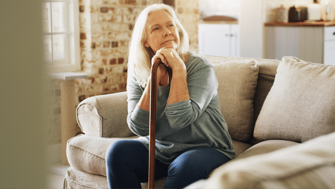 A woman sits on a couch in her home, thoughtfully gazing to the right. She is leaning on her cane.