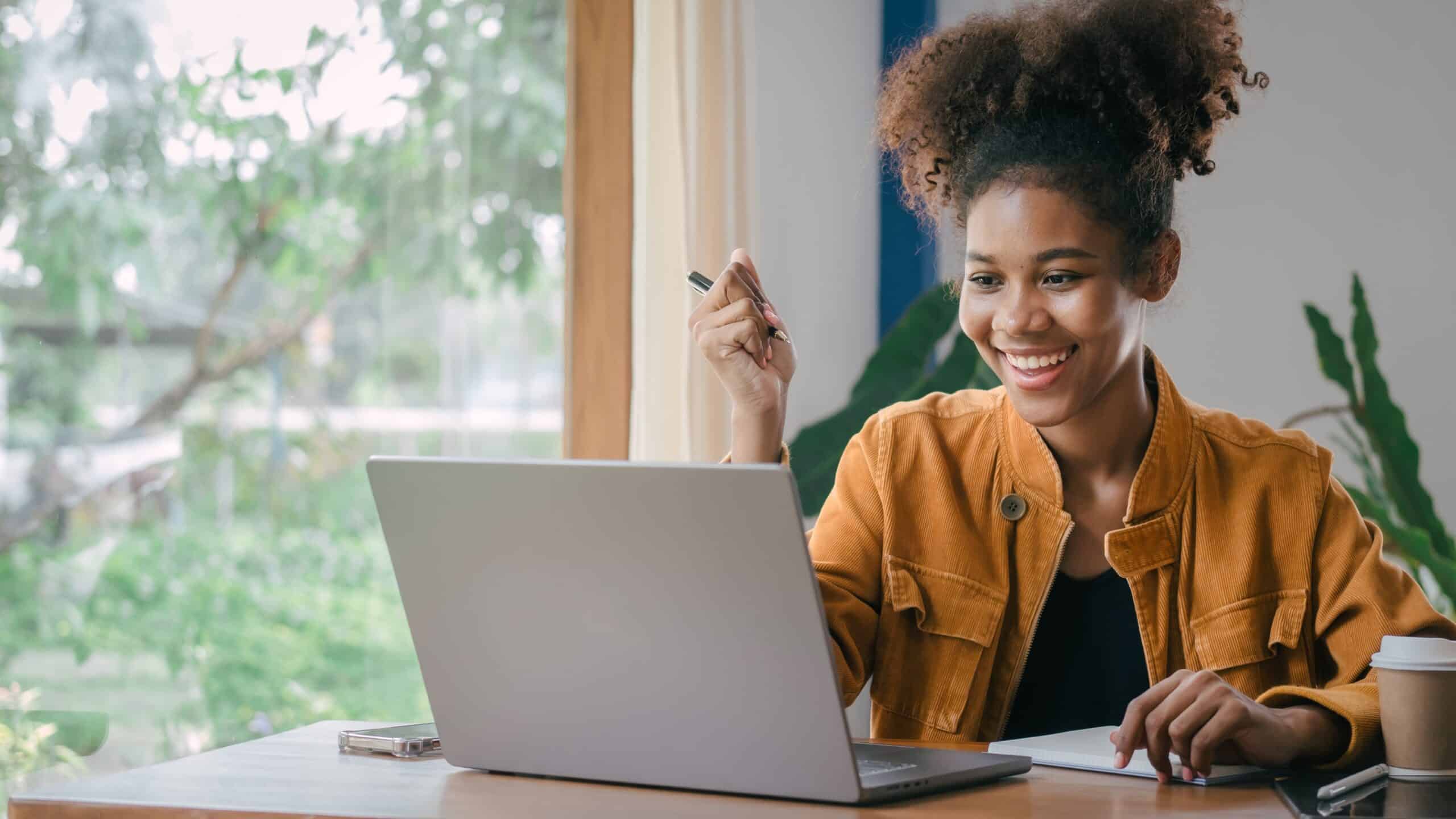 A young black woman holding a pen smiles while looking at a computer screen.