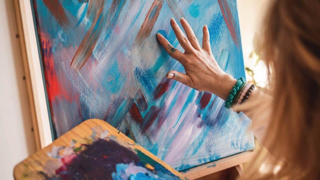A woman wearing bracelets fingerpaints blue and purple on a canvas. No faces are shown.