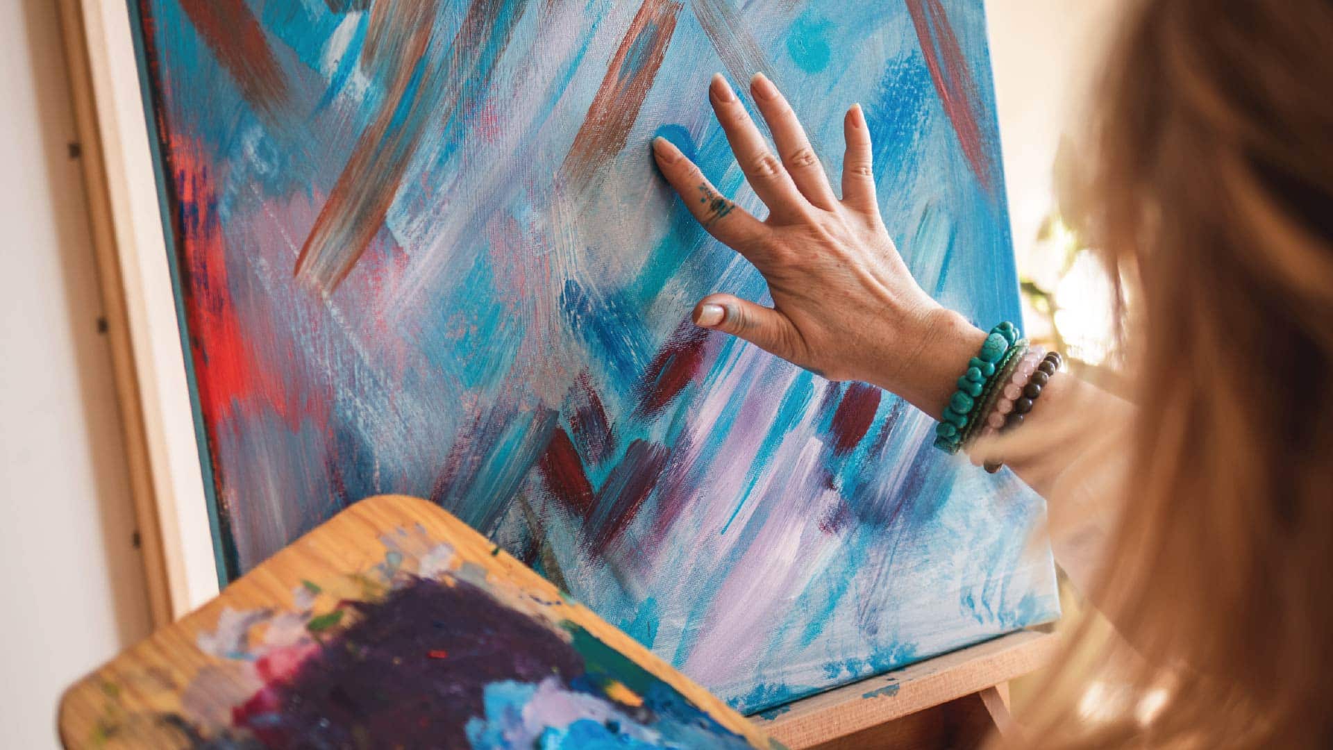 A woman wearing bracelets fingerpaints blue and purple on a canvas. No faces are shown.